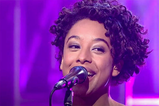 Close-up of a young woman with curly hair smiling and singing into a microphone under purple stage lighting.