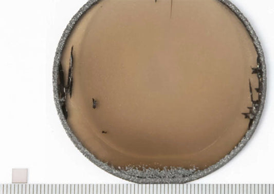 Close-up of a damaged optical lens with visible cracks and chips along the edges. The lens is shown with a ruler for scale, providing detailed insight into the imperfections and texture.