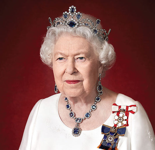 Portrait of Queen Elizabeth II wearing a royal tiara adorned with diamonds and sapphires, matching earrings, and an elegant necklace. She is also wearing official royal decorations, including a ribbon and medal, against a red background.
