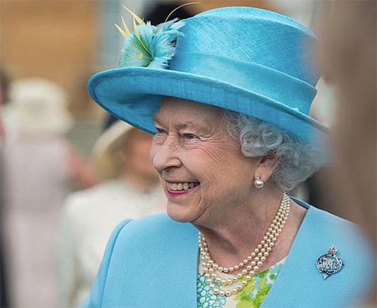 Queen Elizabeth II smiling and wearing a light blue hat, blue jacket, pearl necklace, and brooch, with a cheerful expression in a public appearance.