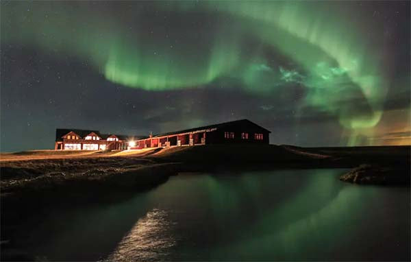 Stunning view of the Northern Lights over a rustic lodge with a reflection in the calm water, showcasing the beauty of nature in a nighttime landscape.