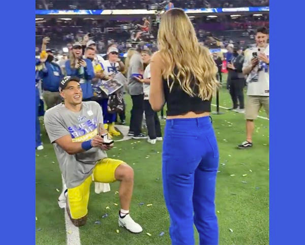 Man proposing with an engagement ring on the football field during the Super Bowl celebration. The woman stands in front of him, smiling, while fans cheer in the background.