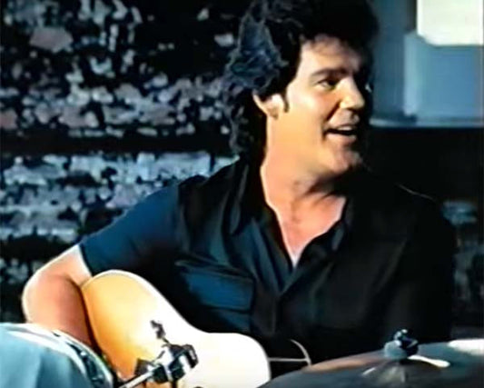 Man with dark hair smiling and playing an acoustic guitar, sitting next to a drum set in a music studio with a brick wall background.