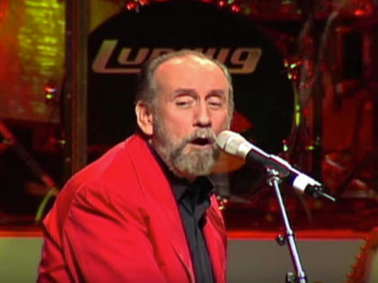 Legendary musician performing on stage, wearing a red jacket, singing into a microphone with a drum kit in the background.