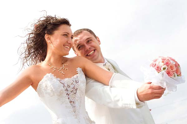 Happy newlywed couple in wedding attire, the bride holding a bouquet of pink roses and smiling at her groom in a romantic outdoor setting with blue skies.