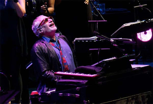 Musician playing the piano passionately on stage while wearing sunglasses and a leather jacket.