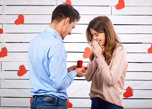 Couple engaged in a romantic proposal with the man presenting a red ring box to the woman surrounded by heart decorations.