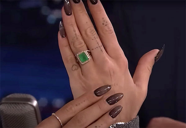 Close-up of a hand with several tattoos and dark manicured nails, wearing a statement ring with a green gemstone, likely emerald, on the ring finger.