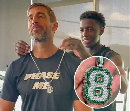 Football player Aaron Rodgers smiling while his teammate helps him wear a custom jewelry necklace featuring a diamond '8' pendant.