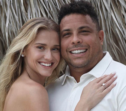 Ronaldo Nazário, the famous Brazilian footballer, smiles alongside his fiancée, showing off her engagement ring in a beachside photo. They both look happy and relaxed.