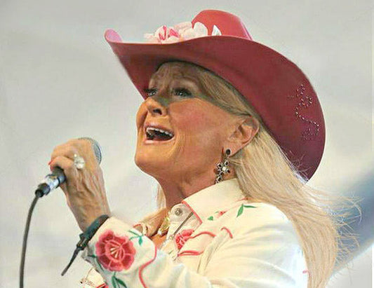 Female country singer wearing a pink cowboy hat and floral western attire, singing passionately into a microphone during a live performance.