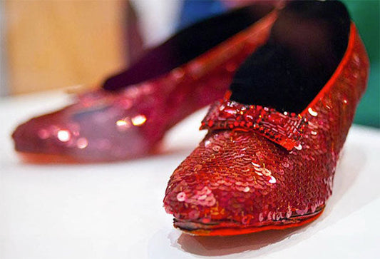 Close-up image of red sequin Dorothy-style shoes, featuring a bow on the toe, displayed on a white surface.