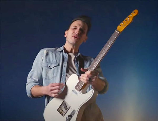 A male musician playing a white electric guitar while performing on stage, wearing a denim jacket with a blue gradient background, singing passionately.