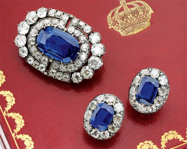Elegant vintage jewelry set featuring a blue sapphire brooch with diamond accents, paired with matching blue sapphire earrings, displayed on a red background with a golden crown motif.