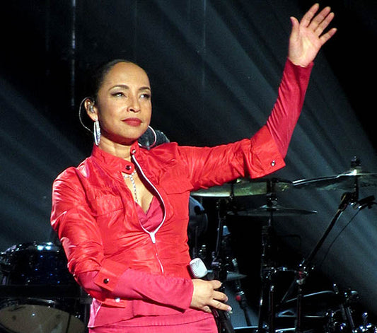 Sade Adu performing on stage in a red outfit, waving to the audience during a live concert with a band in the background.