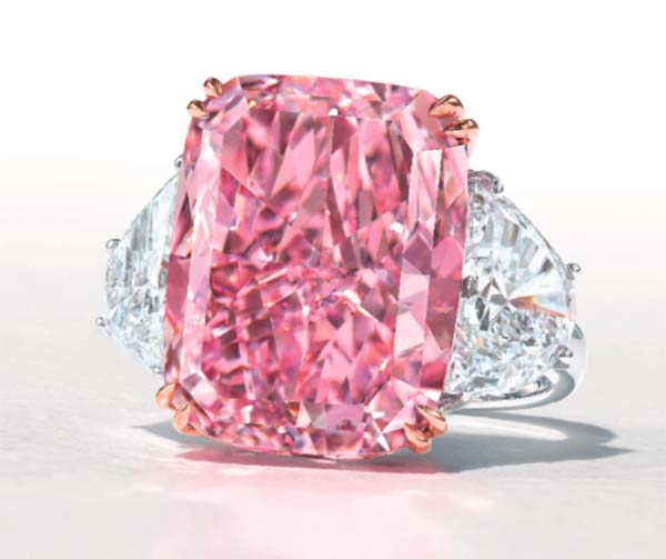 Large pink cushion-cut diamond ring with two white trapezoid diamonds on either side, set in a platinum band with rose gold accents.