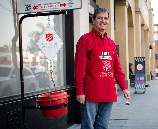 Salvation Army volunteer wearing a red apron while standing next to a red kettle for donations on a street, promoting the organization's cause.