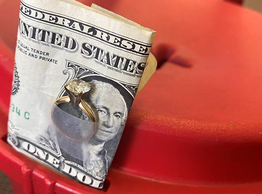 A close-up image of a diamond engagement ring on a dollar bill, placed on a red surface, symbolizing love and commitment.