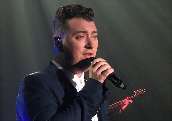 Sam Smith performing live on stage, singing into a microphone while wearing a dark suit and a white shirt. The stage lights create a dramatic effect, highlighting their expression and vocals during the performance.