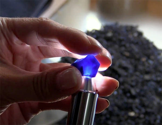 Close-up of a hand holding a blue gemstone under a light source, with a dark background of unprocessed rough minerals, showcasing the process of gem identification or examination.