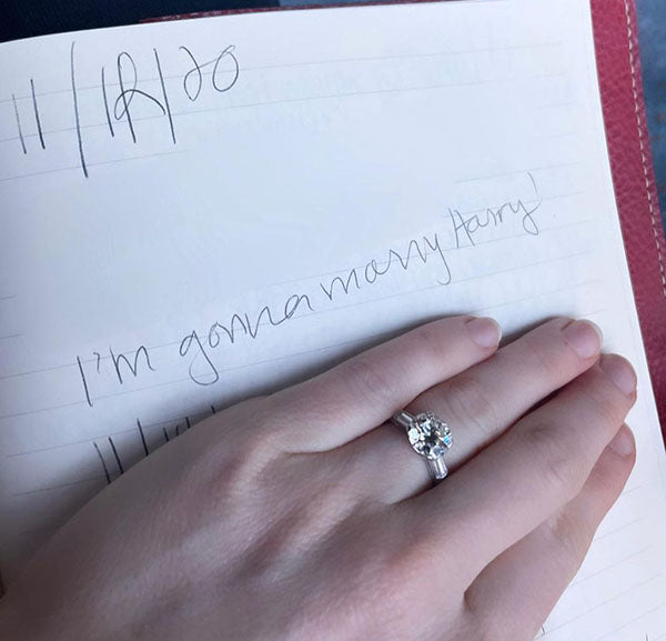 Close-up of a hand wearing a sparkling engagement ring with a round diamond, resting on a notebook with the handwritten note 'I'm gonna marry Harry!' visible on the page.