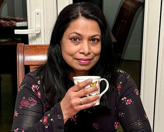 Woman with long dark hair, wearing a floral dress, holding a cup of tea or coffee, sitting at a table in a cozy room.