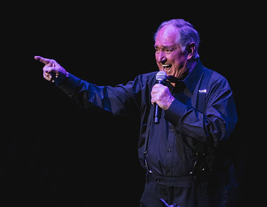 Legendary comedian performing on stage, holding a microphone and pointing while delivering a joke during a live comedy show.