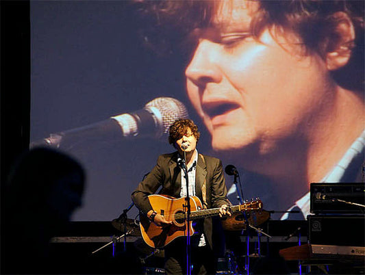 Musician performing on stage with an acoustic guitar, captured in front of a large screen displaying a close-up of his face as he sings into a microphone. The image highlights the live performance and the musician's expression.