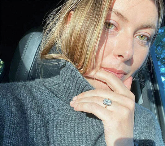 Close-up selfie of a woman wearing a gray turtleneck sweater, with her right hand resting near her chin and showcasing an elegant engagement ring with a large gemstone.