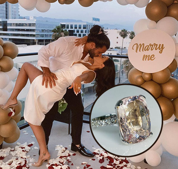 Couple celebrating an engagement with a kiss under a 'Marry Me' balloon arch, with a close-up of the engagement ring showing a radiant-cut diamond on a silver band.