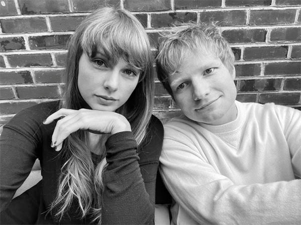 Black and white photo of Taylor Swift and Ed Sheeran sitting together against a brick wall, smiling and posing for the camera. Taylor has long bangs and Ed is wearing a white sweater.