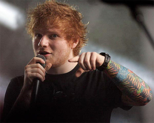 Ed Sheeran performing on stage with a microphone, pointing towards the camera, wearing a black t-shirt, showcasing his colorful sleeve tattoos.