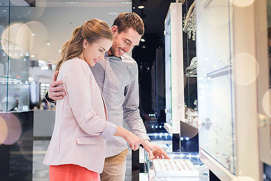 A couple shopping for jewelry in a store, smiling as they point at a jewelry display case with sparkling rings, surrounded by a bright and elegant shopping environment.