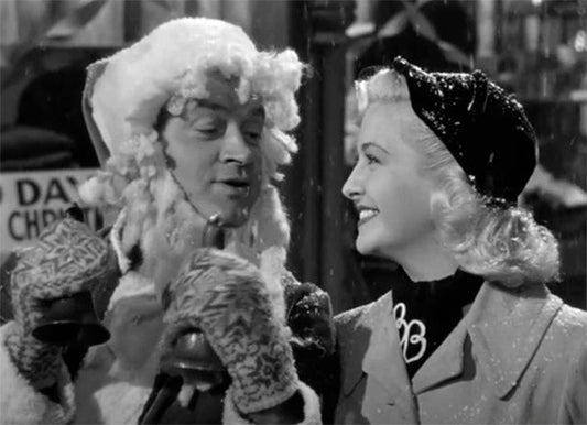 Black-and-white image of a man dressed as Santa Claus and a woman smiling at each other while holding bells, in a snowy outdoor Christmas setting.