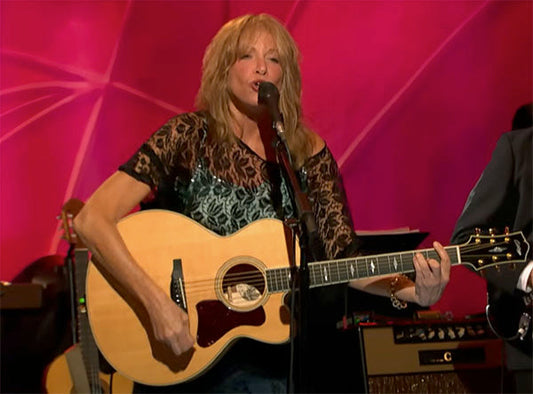 Female musician performing live, playing acoustic guitar while singing into a microphone, with a vibrant stage background.