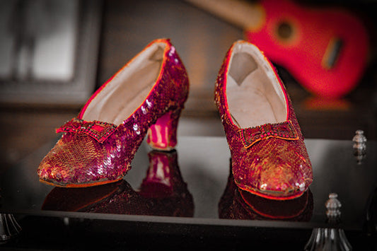 Close-up of a pair of sparkling red sequin shoes with small heels, displayed on a reflective surface with a blurred background.