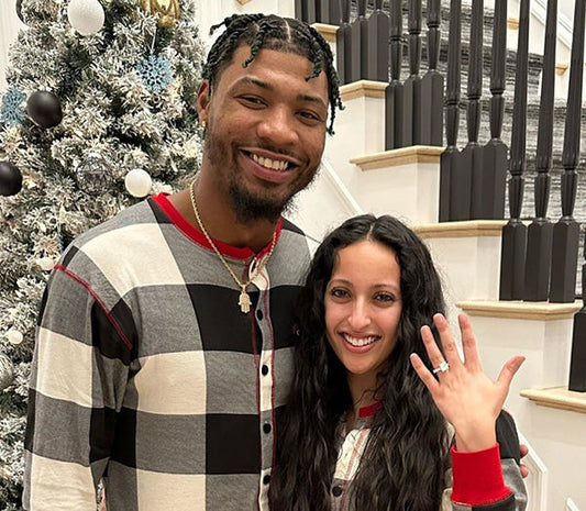 Happy couple celebrating their engagement in front of a Christmas tree, with the woman showing off her engagement ring, wearing matching holiday pajamas.