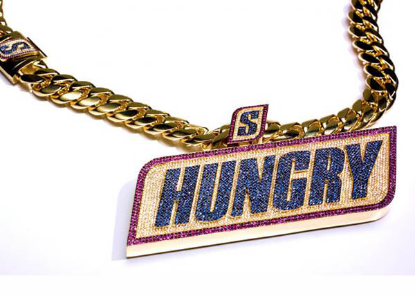 Close-up of a gold chain necklace featuring a large, jeweled pendant with the word 'HUNGRY' in bold letters. The pendant is adorned with colorful gemstones, highlighting the stylish and flashy design of the piece.
