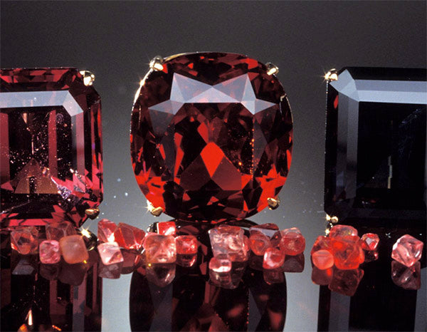 Close-up of deep red garnet gemstones in various cuts and sizes, including raw and faceted stones, displayed on a reflective surface.