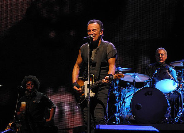 Bruce Springsteen performing on stage with his guitar, singing into a microphone, while his band plays in the background at a live concert.