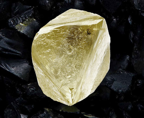 Close-up of a yellow diamond crystal with sharp edges, set against a dark, textured background of coal or charcoal, highlighting its unique shape and color.