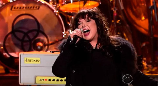 Female rock singer passionately performing on stage with a microphone, Led Zeppelin drum set and guitar amp in the background during a live concert tribute.