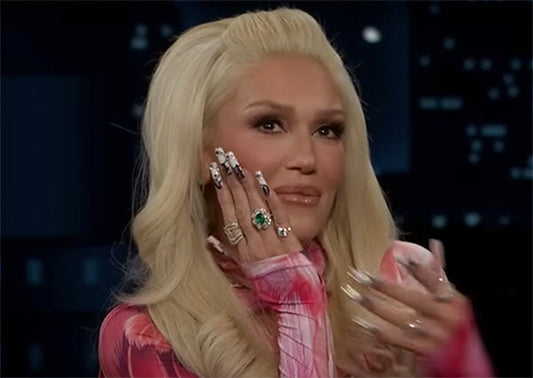 Gwen Stefani smiling with her hand to her face, showcasing her glamorous nails adorned with various rings, including an eye-catching green stone ring, on a late-night talk show.