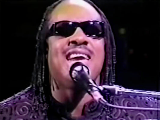 Stevie Wonder performing on stage, wearing sunglasses, singing passionately into a microphone while playing the piano.