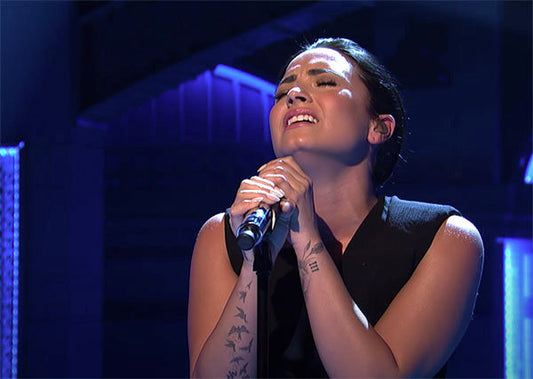 Demi Lovato performing live on stage, singing passionately with a microphone in hand, expressing emotion through music, with visible tattoos on her arms.