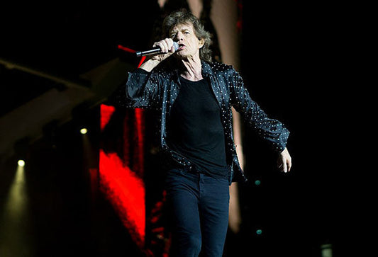 Mick Jagger of The Rolling Stones performing on stage with a microphone, wearing a black shirt with silver sparkles.