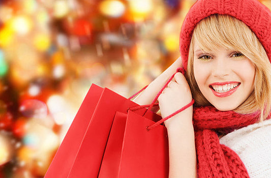 Smiling woman wearing a red knitted hat and scarf, holding shopping bags with a colorful bokeh Christmas background.