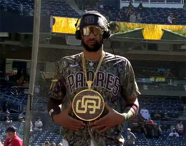 Baseball player wearing a Padres camouflage jersey, gold chain, and headphones, holding a large gold belt with the Padres logo at a game.