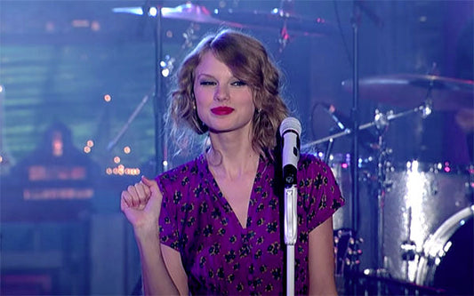 Taylor Swift performing on stage with a microphone, wearing a purple floral dress and a confident smile.
