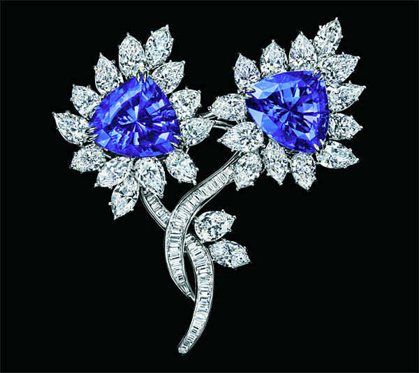 A luxurious diamond brooch featuring two flower-shaped designs with purple sapphire center stones surrounded by diamond petals, set in silver.